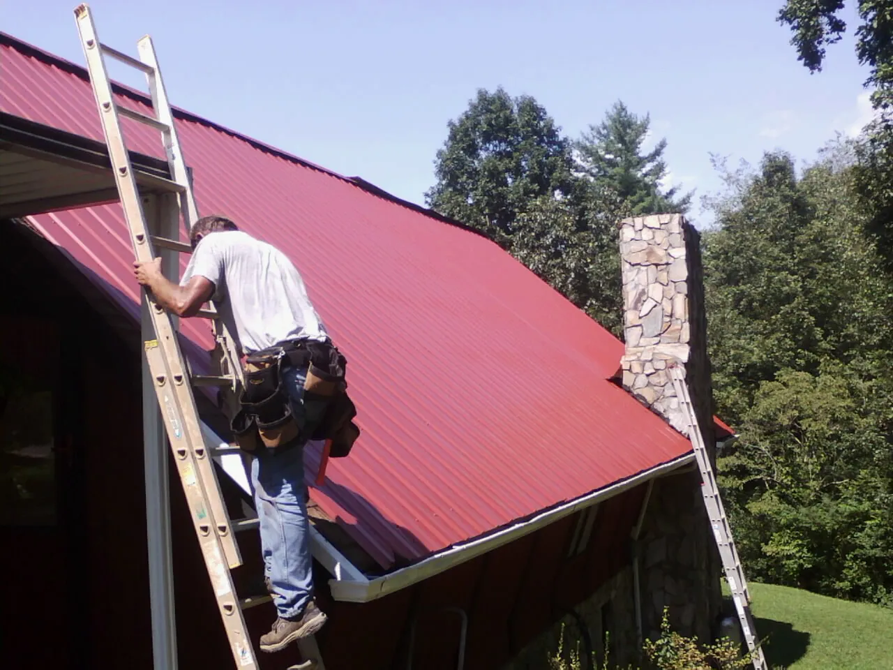 Roofer on a ladder beside a red metal roof and stone chimney — Soffit & Fascia Repair by Roofing Brothers in Edgemoor